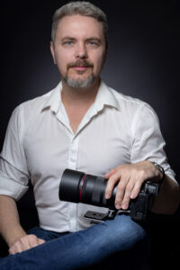 Headshot photograph of Laszlo Kupi sitting down, holding a camera
