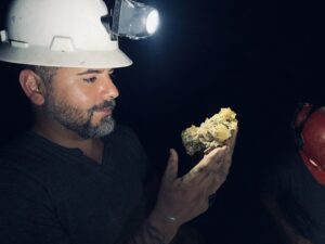 Profile of Daniel Ulibarri examining and holding a mineral specimen in a mine while wearing a hard hat with a headlamp brightly shinning on the specimen